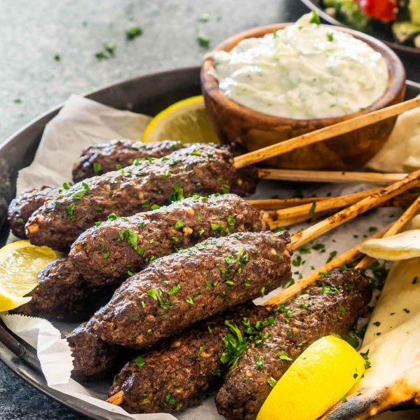 a few beef koftas on a plate with some pitas garnished with parsley next to tzatziki sauce.
