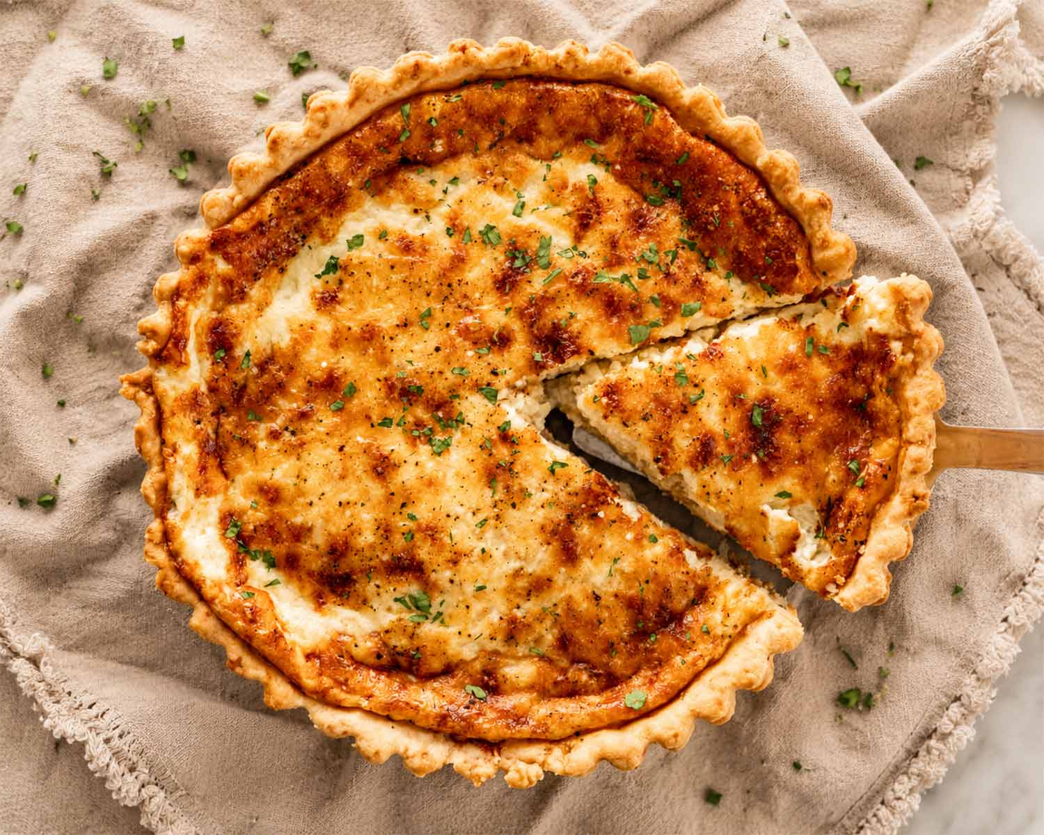 Overhead view of golden Quiche Lorraine on a beige linen cloth with one slice lifted, topped with herbs and crispy browned cheese.