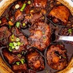 Close up of braised oxtail in rich dark gravy, spoon lifting tender piece showing glossy sauce and soft beans.