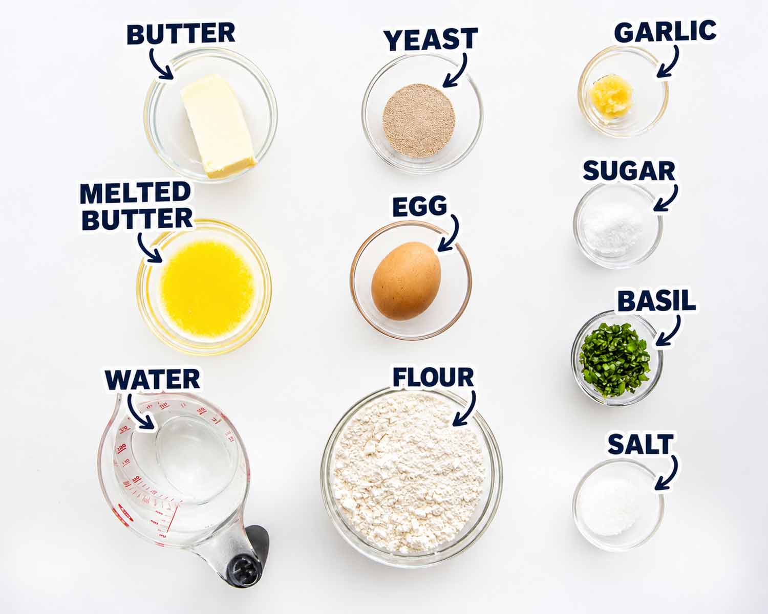 Labeled garlic knot ingredients, including flour, yeast, butter, egg, garlic, basil, sugar, salt, and water, arranged on a white surface.