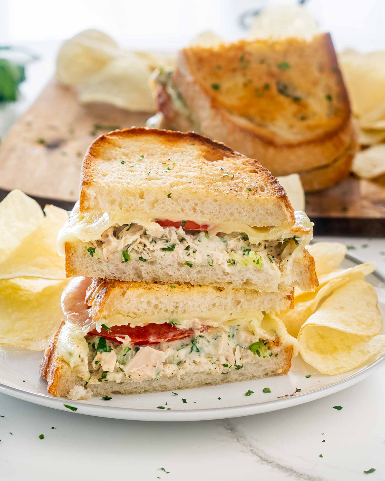 a tuna melt with tomatoes, cut in two and stacked on a white plate with potato chips.