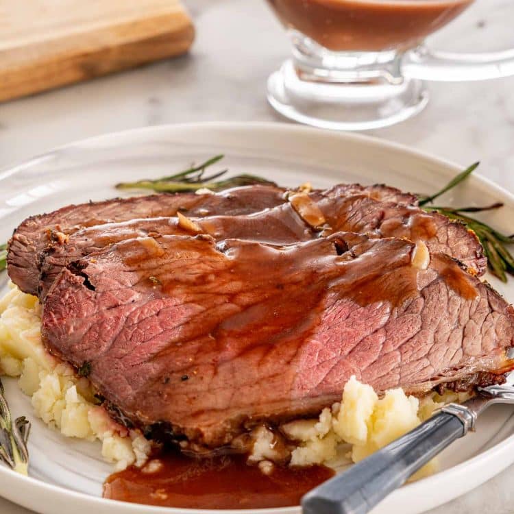 Angled closeup of roast beef slices over mashed potatoes with brown gravy, rosemary, and a gravy boat in the background.