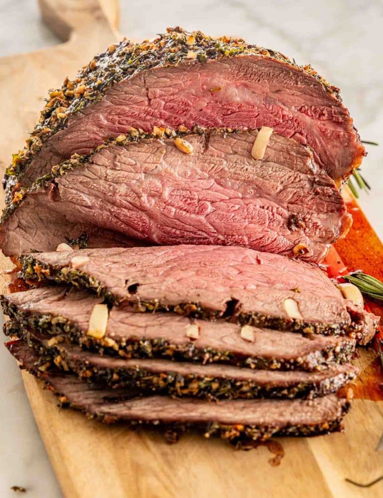 Closeup of garlic-studded roast beef with a browned herb crust and pink slices fanned across a wooden cutting board.