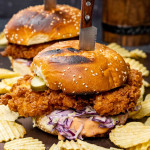a crispy fried chicken sandwich with coleslaw on a cutting board with potato chips.