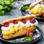 a gorgeous eclair filled with vanilla custard cream and topped with whipped cream and raspberries on a plate.