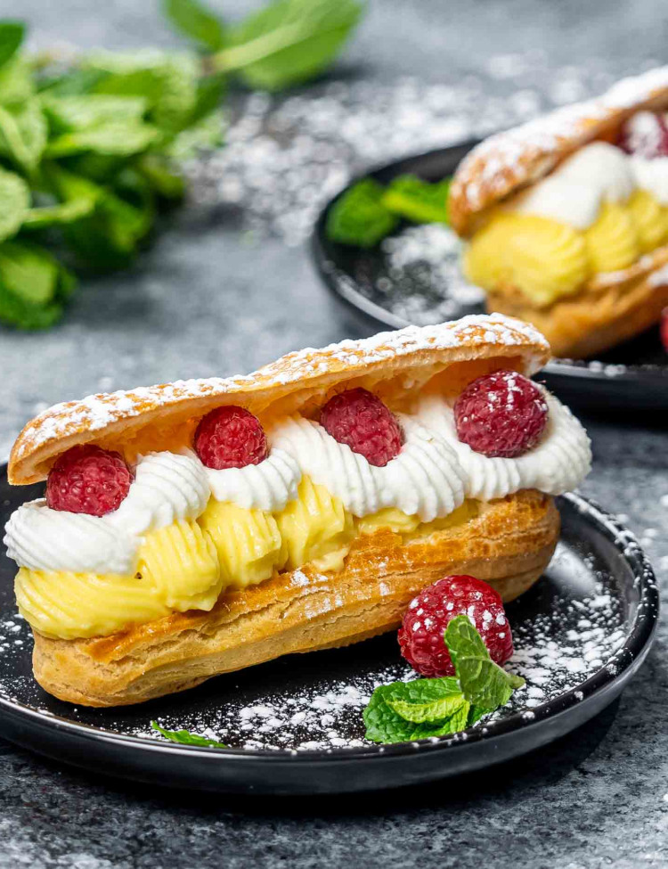 a gorgeous eclair filled with vanilla custard cream and topped with whipped cream and raspberries on a plate.