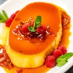 a gorgeous creamy flan on a white oval plate garnished with raspberries.