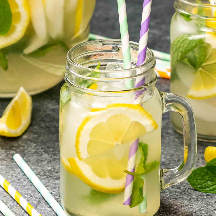 a jar full with lemonade, lemon slices, mint and a couple straws with a pitcher of lemonade in the background.