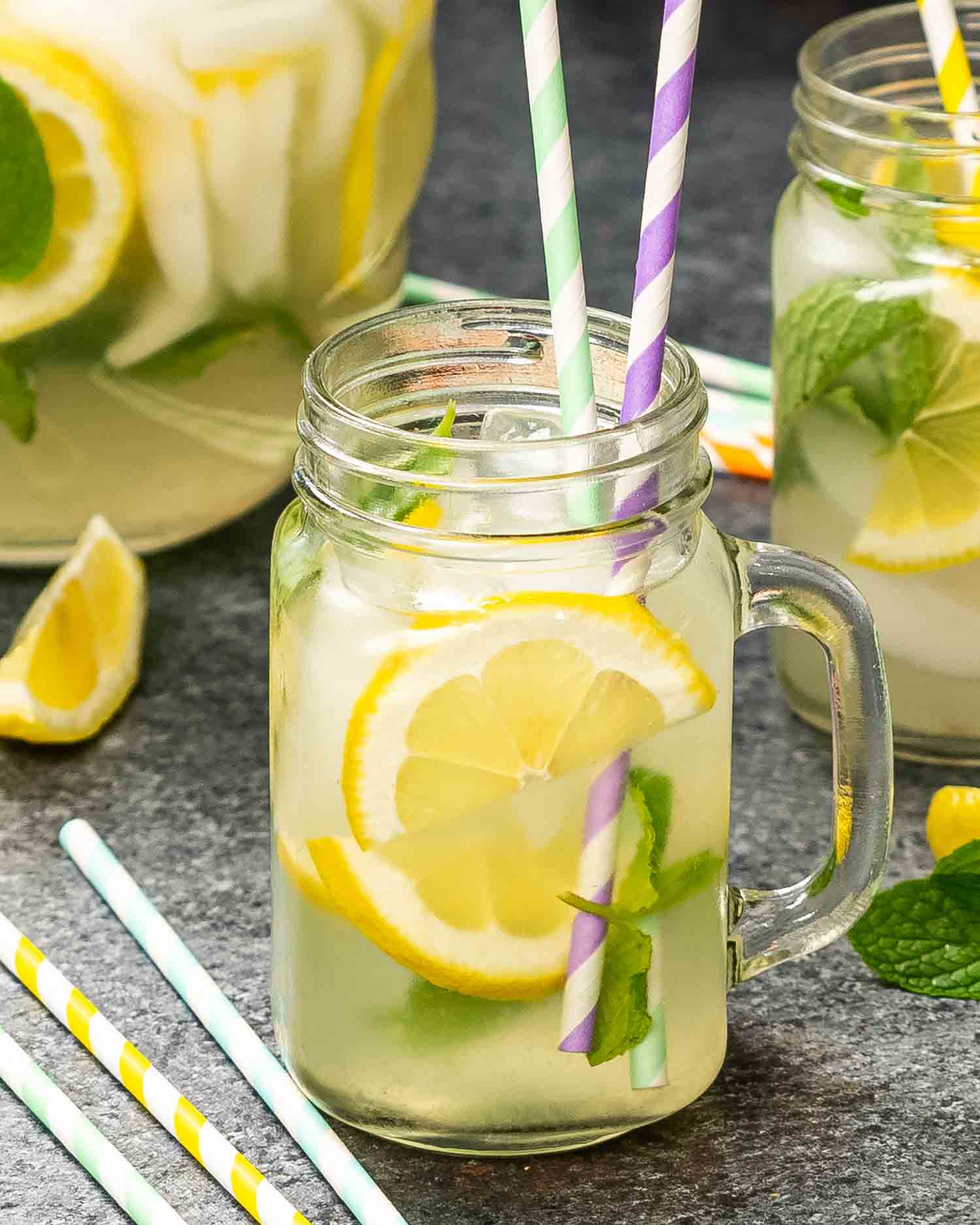 a jar full with lemonade, lemon slices, mint and a couple straws with a pitcher of lemonade in the background.