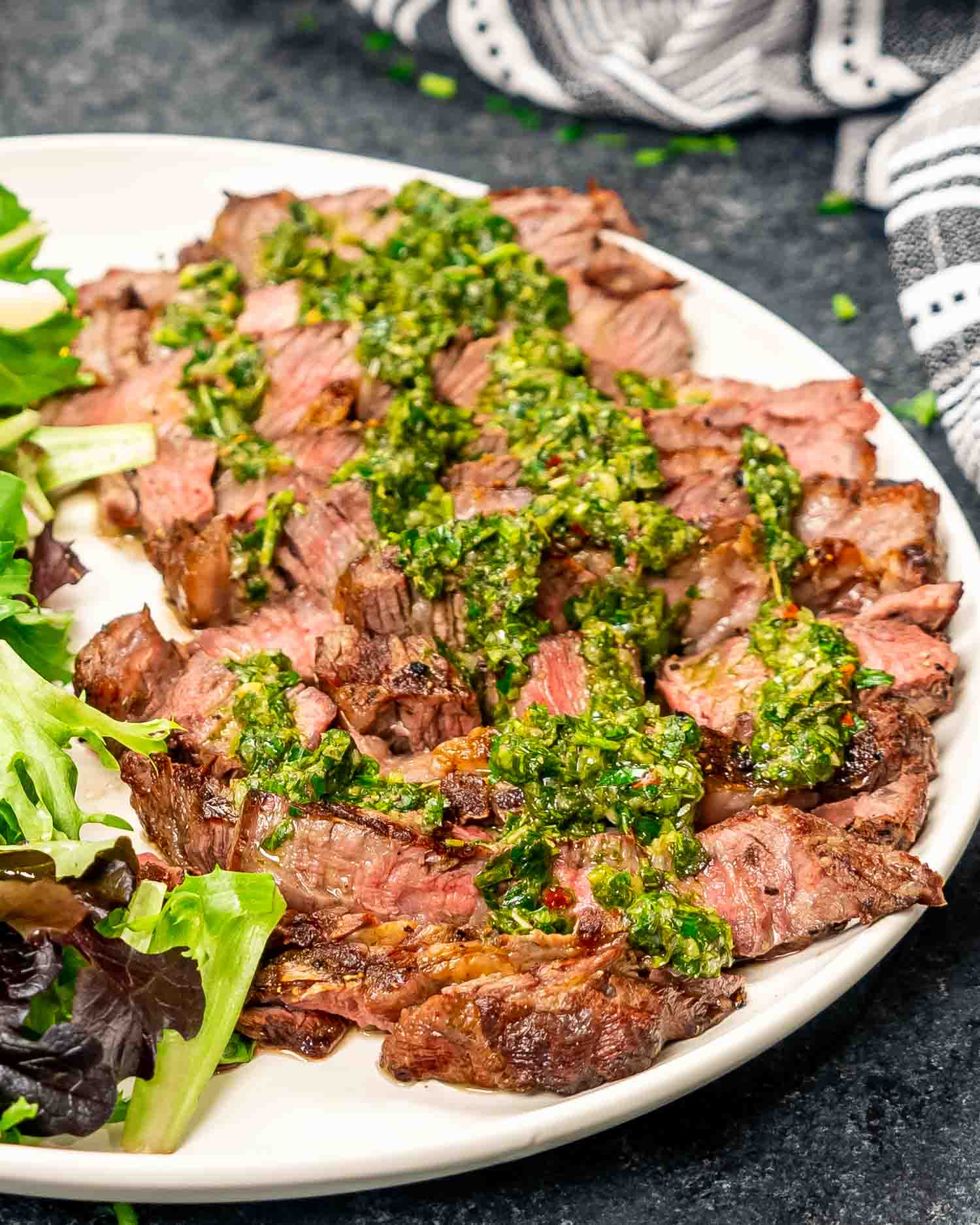 grilled ribeye sliced up drizzled with chimichurri sauce on a white plate along with a tossed green salad.