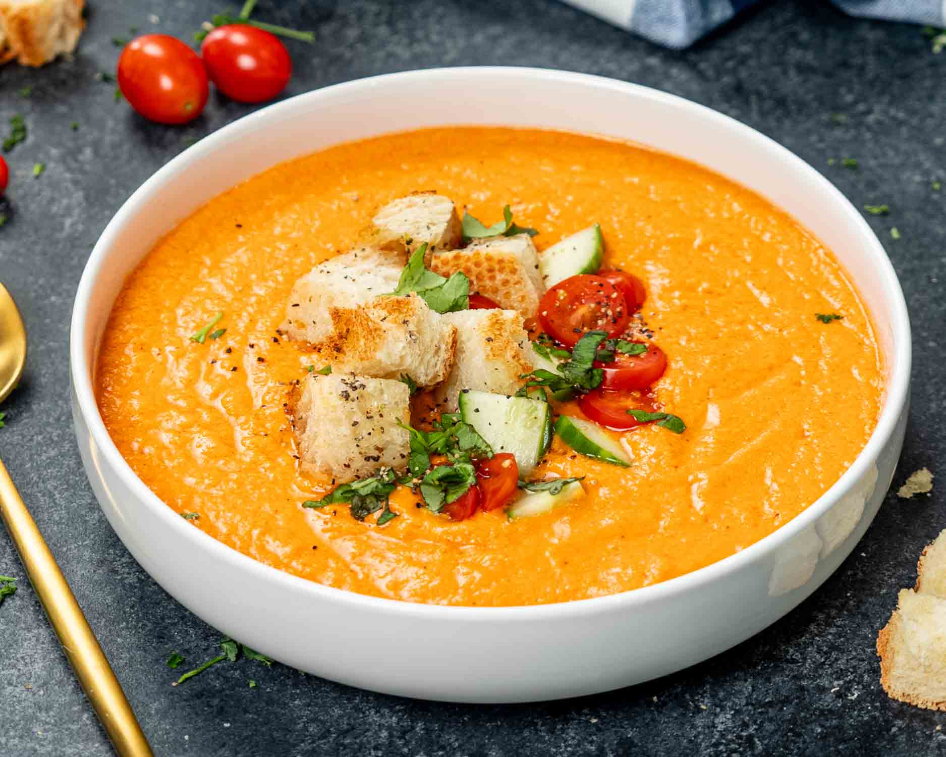 a white bowl loaded with fresh gazpacho and topped with croutons, chopped tomatoes and cucumber and a sprinkle of basil.