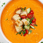 a white bowl loaded with fresh gazpacho and topped with croutons, chopped tomatoes and cucumber and a sprinkle of basil.