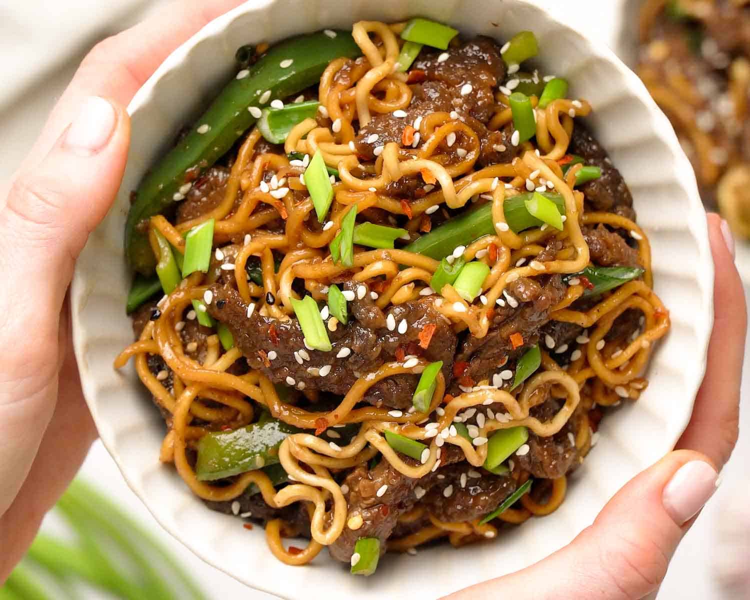 Hands holding a bowl of Mongolian Beef Ramen with sticky sweet savory sauce coating beef and ramen noodles.