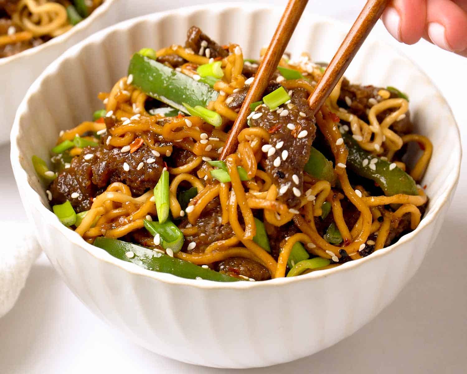 Close up of Mongolian Beef Ramen in a white bowl with chopsticks lifting glossy noodles and tender beef slices.