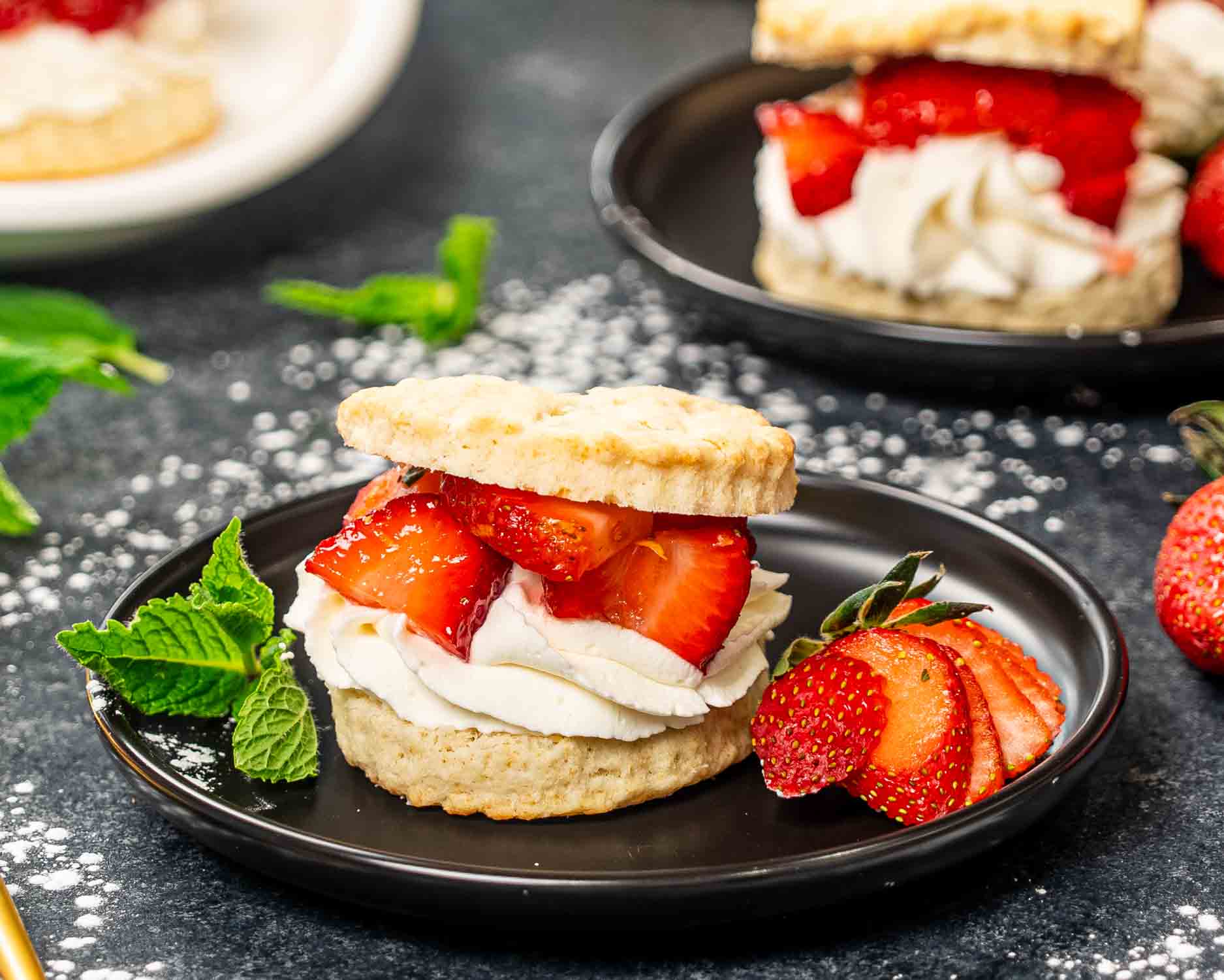 a strawberry shortcake on a black plate with a strawberry on the side and a mint leaf, sprinkled with powdered sugar.