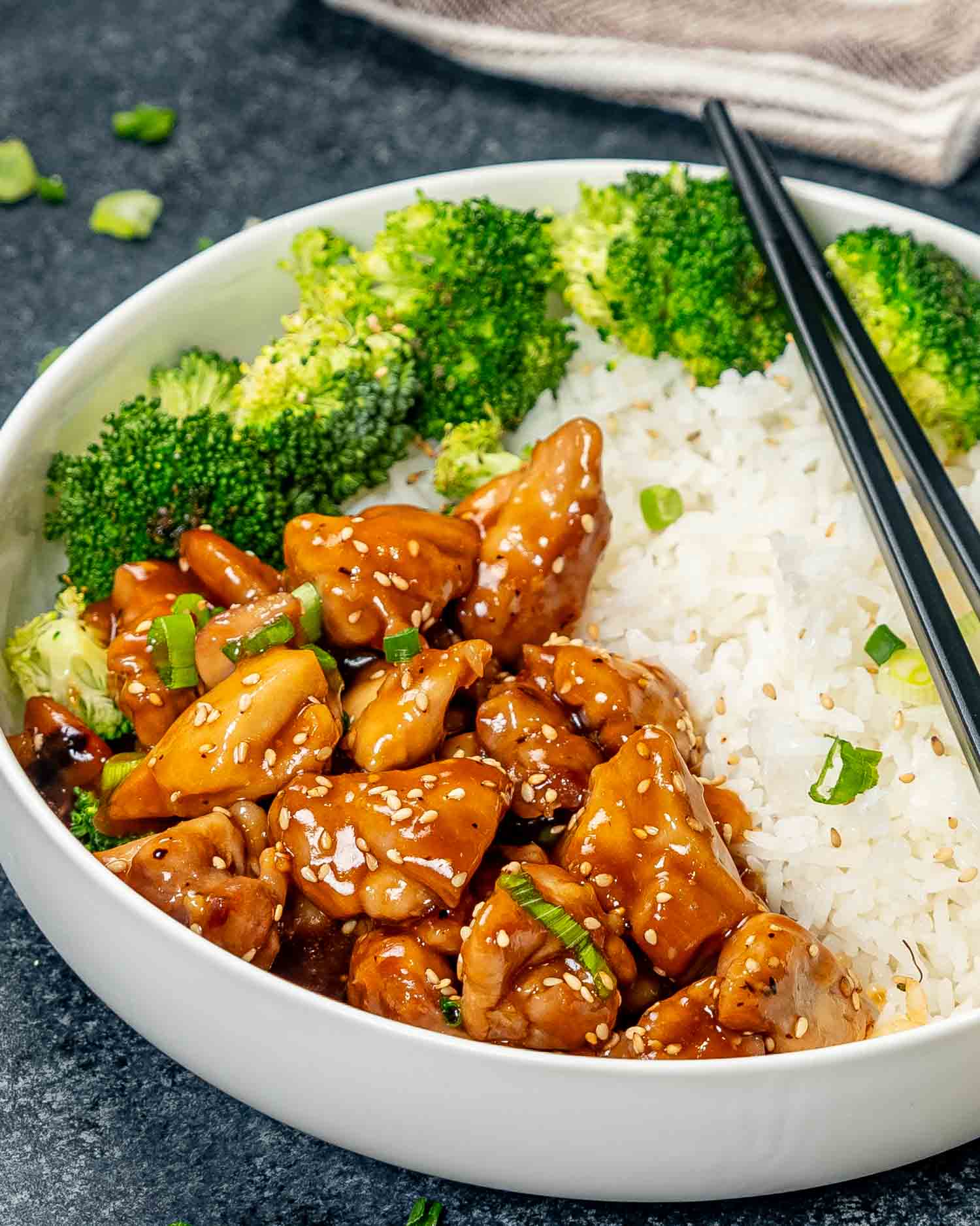 teriyaki chicken in a white bowl with some cooked rice and steamed broccoli.