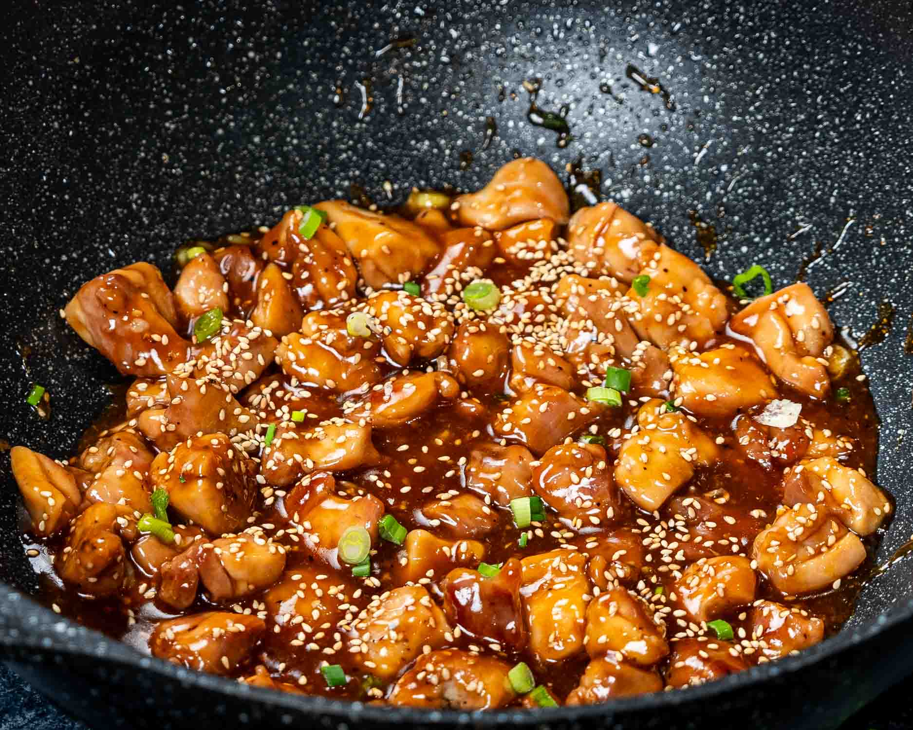 teriyaki chicken garnished with sesame seeds and green onions in a wok.