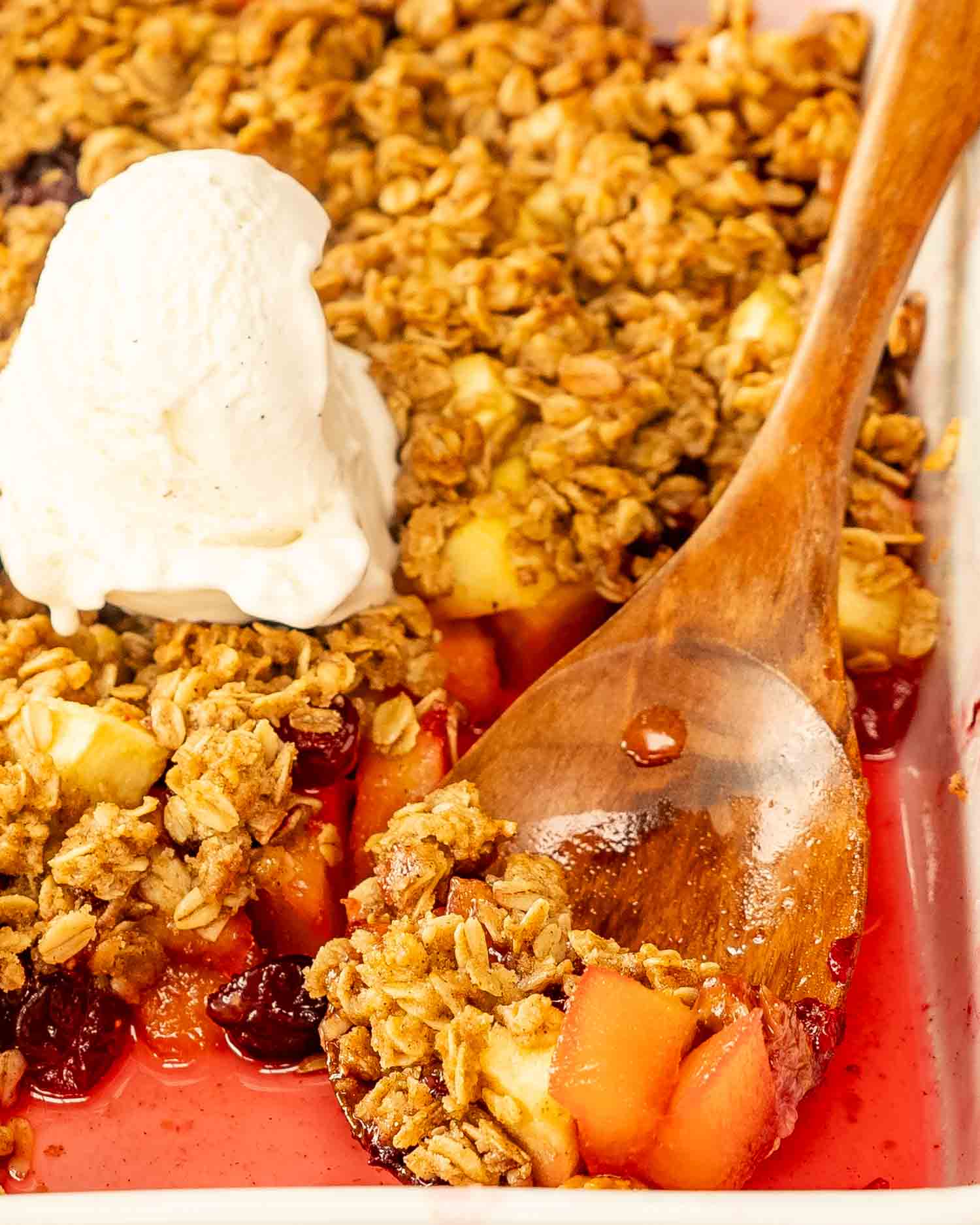 apple cranberry crisp in a casserole dish with a dollop of ice cream in the middle and a serving spoon inside.