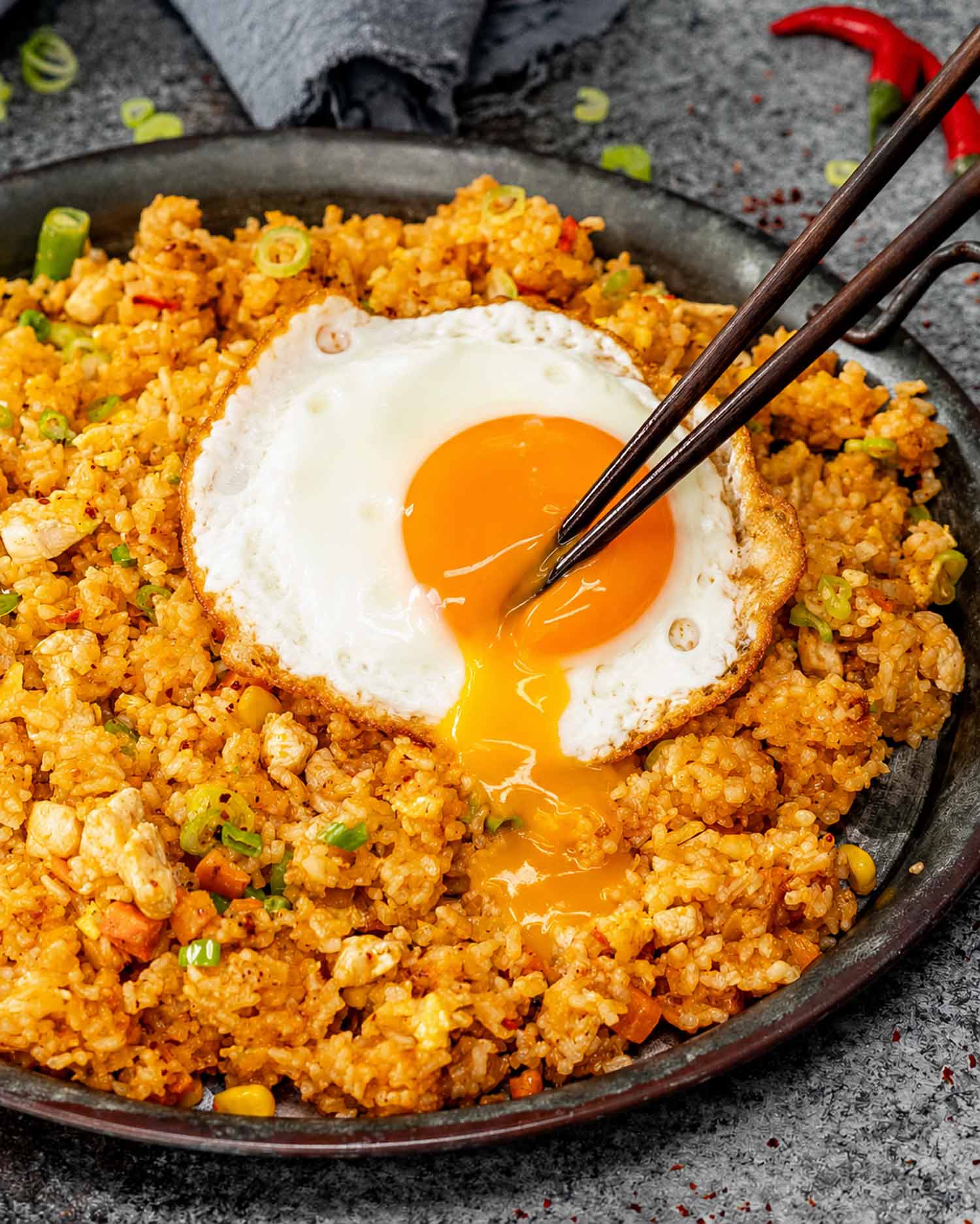 Chopsticks break a glossy fried egg yolk, allowing it to run over chicken nasi goreng in a dark skillet.