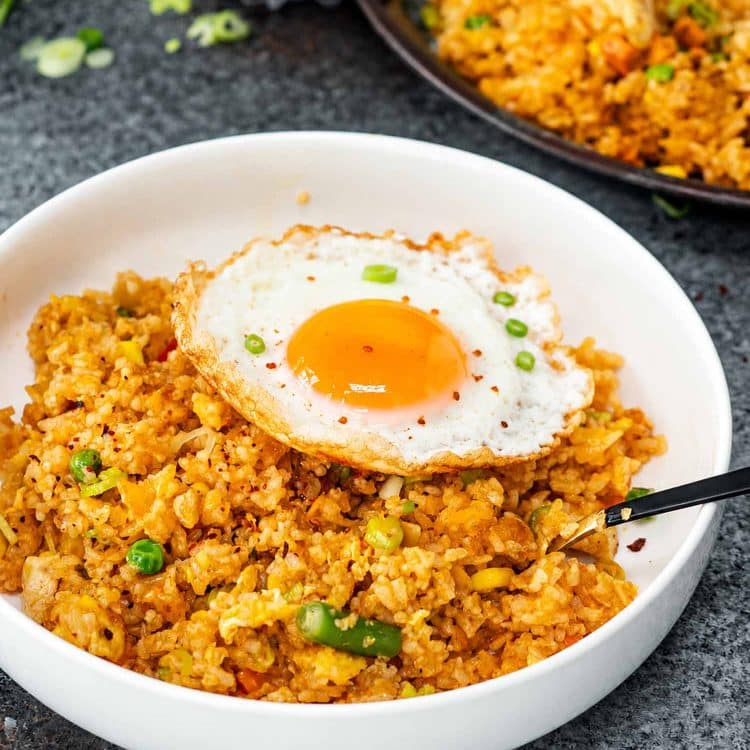 White bowl filled with colorful nasi goreng, topped with a crispy fried egg and garnished with sliced green onions.
