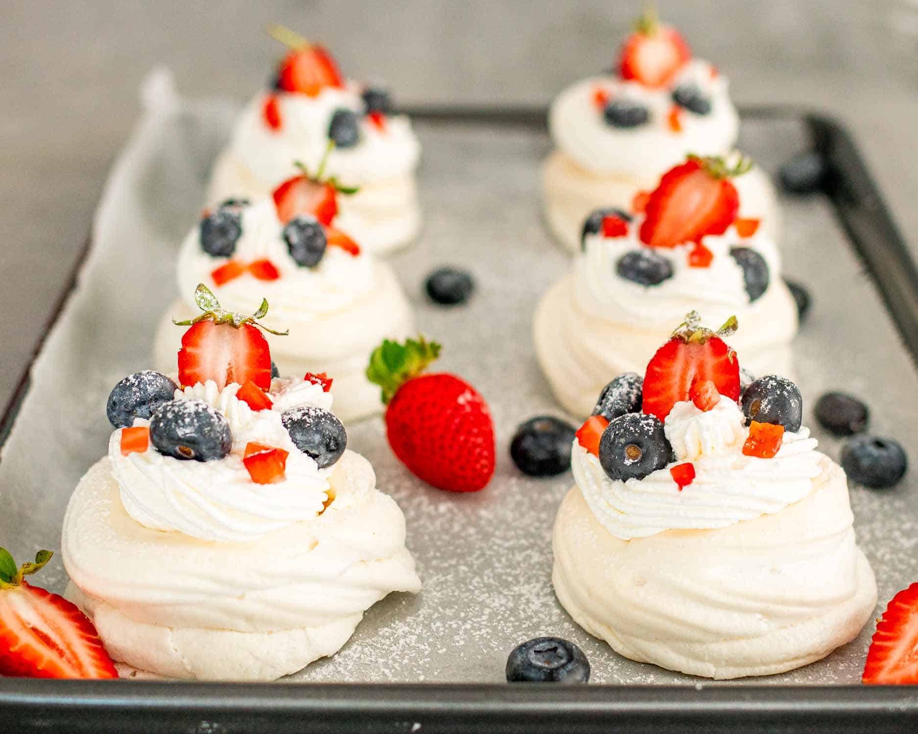 mini pavlovas topped with whipped cream and berries on a baking sheet.