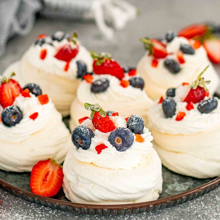 6 mini pavlovas on a metal serving platter topped with whipped cream and fresh fruit.