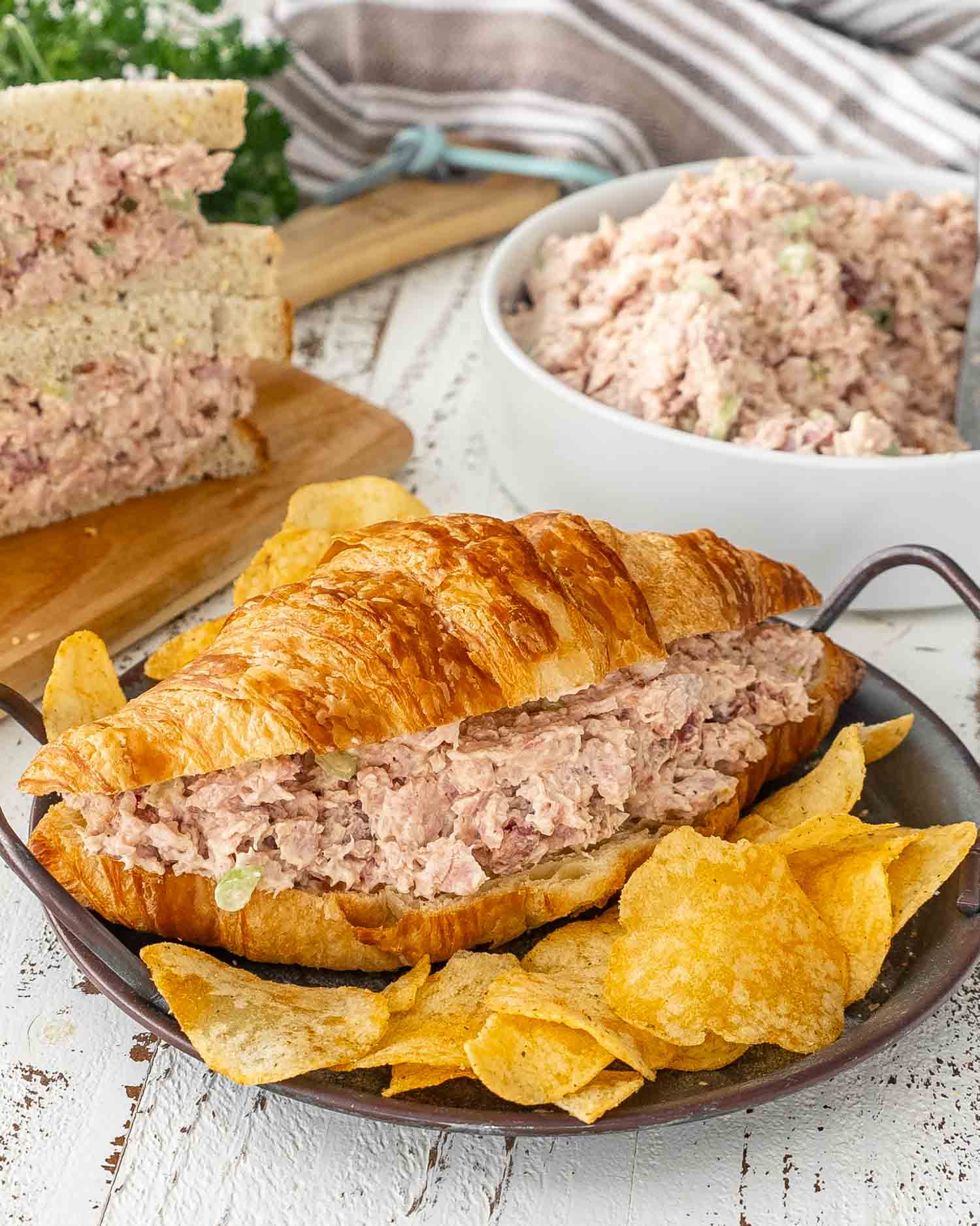 a croissant with ham salad on a plate along side some potato chips.
