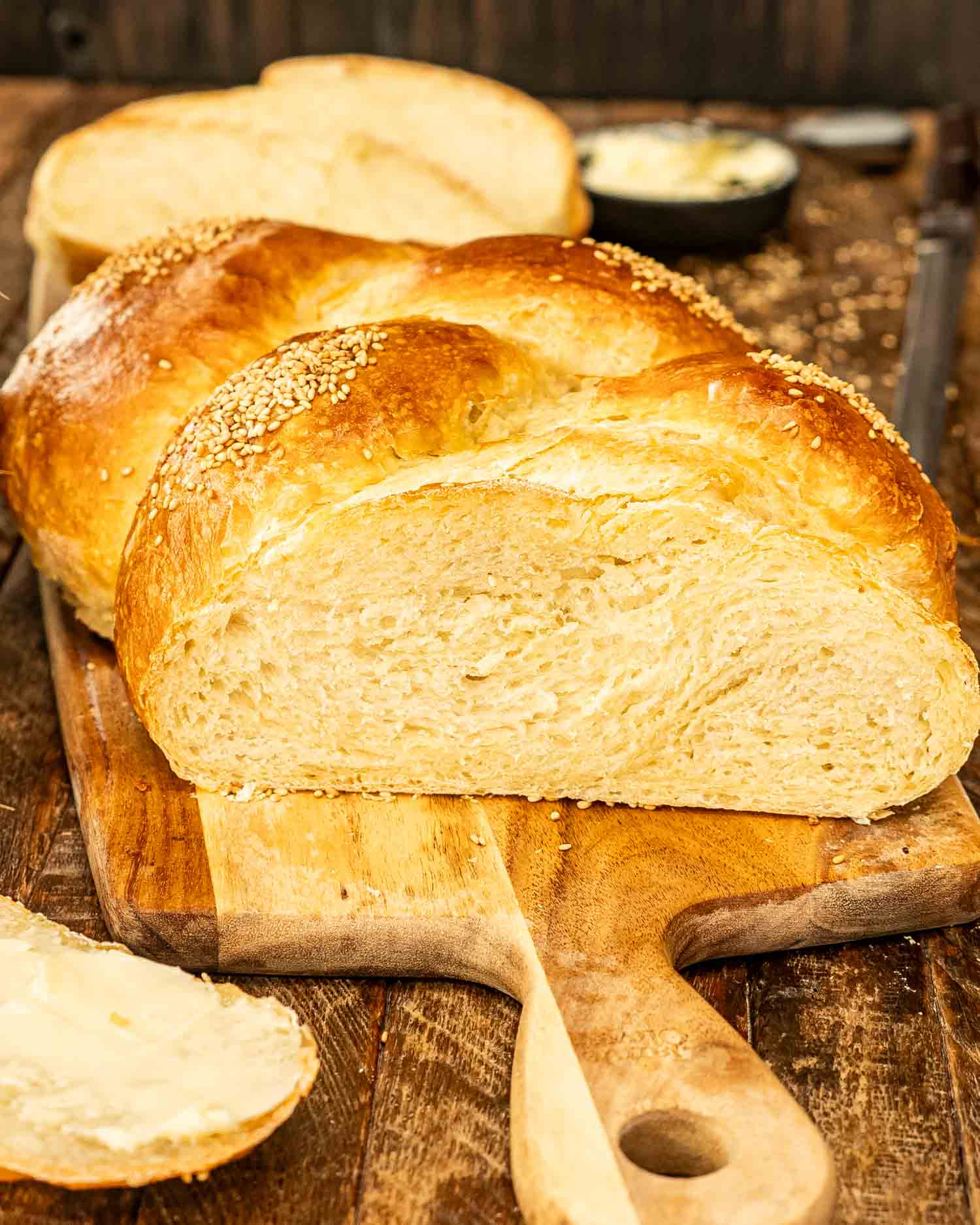 A fresh, golden-braided no knead challah bread on a wooden board with slices and butter nearby.