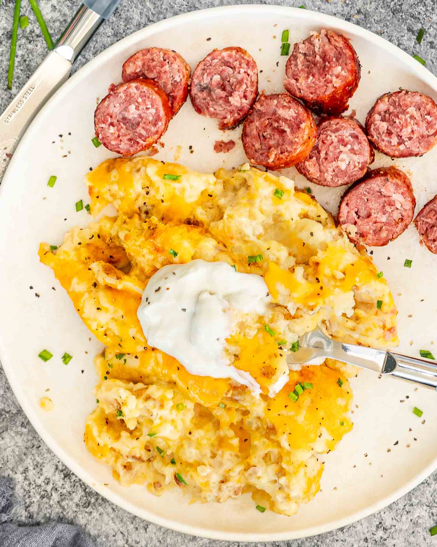 a serving of potatoes romanoff with a dollop of sour cream along some cooked sausage on a white plate.