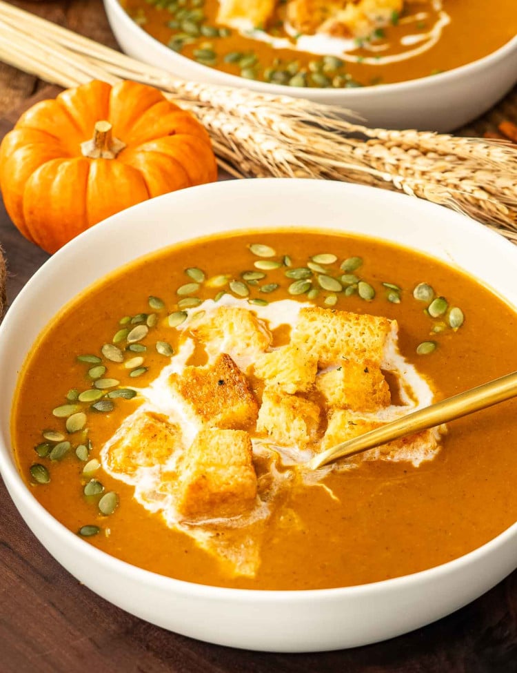 pumpkin soup in a white bowl garnished with pumpkin seeds, croutons, and a drizzle of cream.
