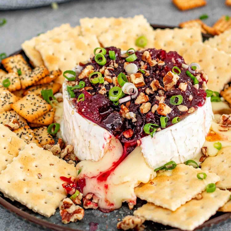 An image showcasing a perfectly baked wheel of Brie cheese topped with a rich, ruby-red cranberry sauce and sprinkled with chopped pecans and sliced green onions. The cheese is oozing out enticingly onto a dark plate, surrounded by an assortment of crackers.