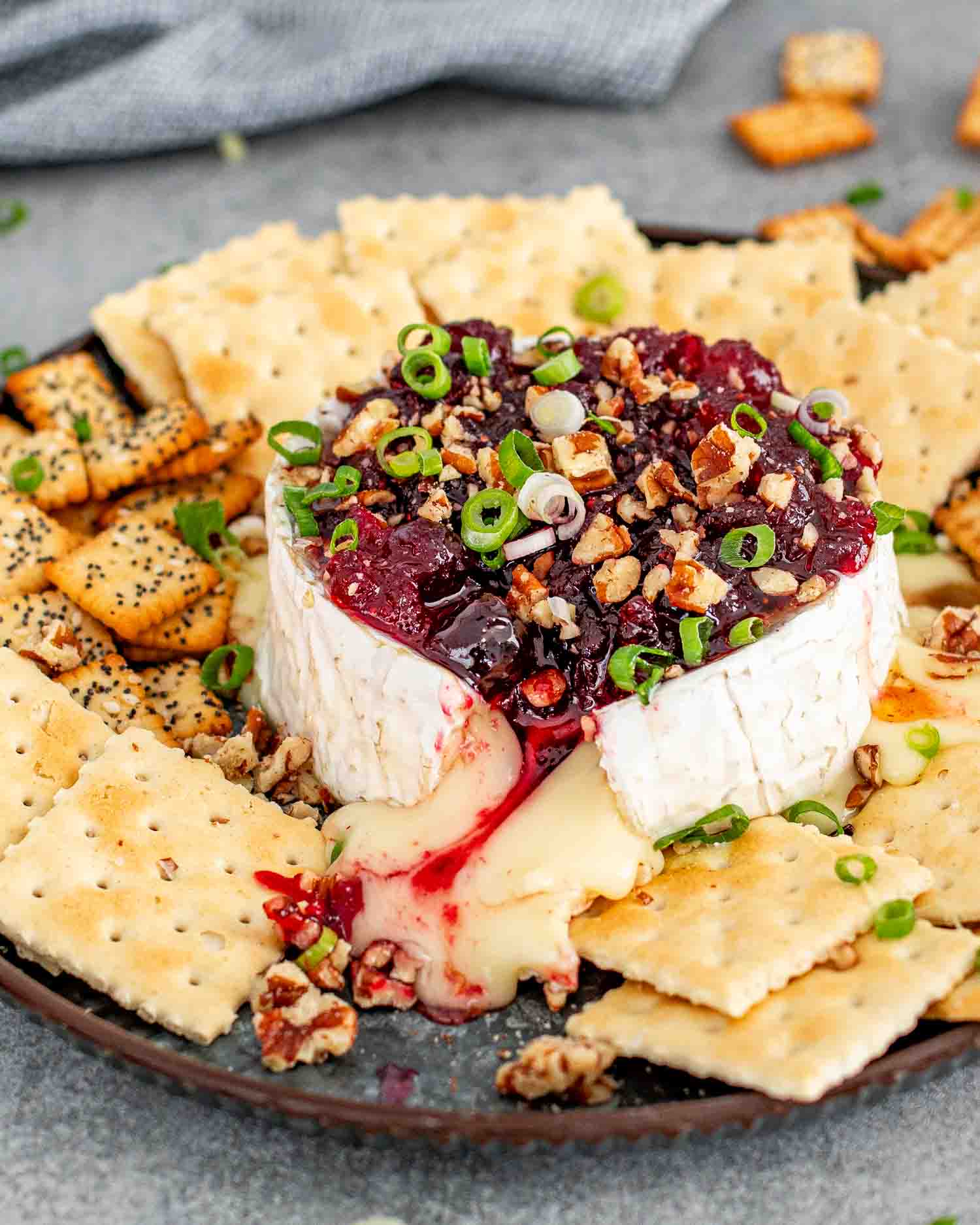An image showcasing a perfectly baked wheel of Brie cheese topped with a rich, ruby-red cranberry sauce and sprinkled with chopped pecans and sliced green onions. The cheese is oozing out enticingly onto a dark plate, surrounded by an assortment of crackers. 