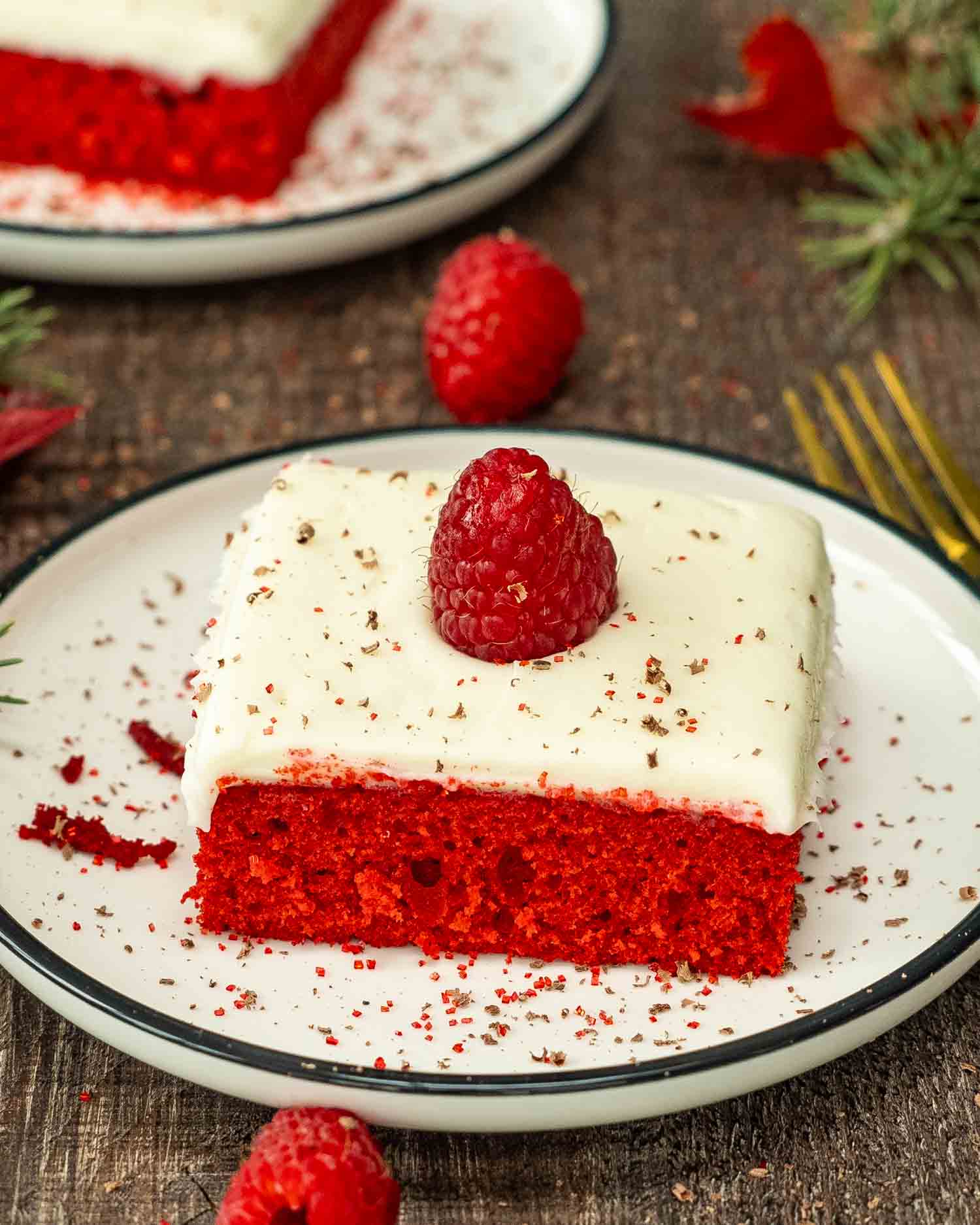 a slice of red velvet sheet cake with cream cheese frosting on a white dessert plate topped with a raspberry.