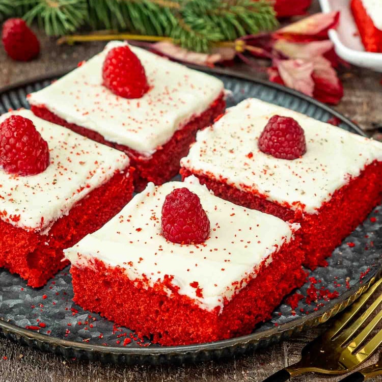 4 slice of red velvet sheet cake topped with cream cheese frosting and each topped with a raspberry on a metal plate.