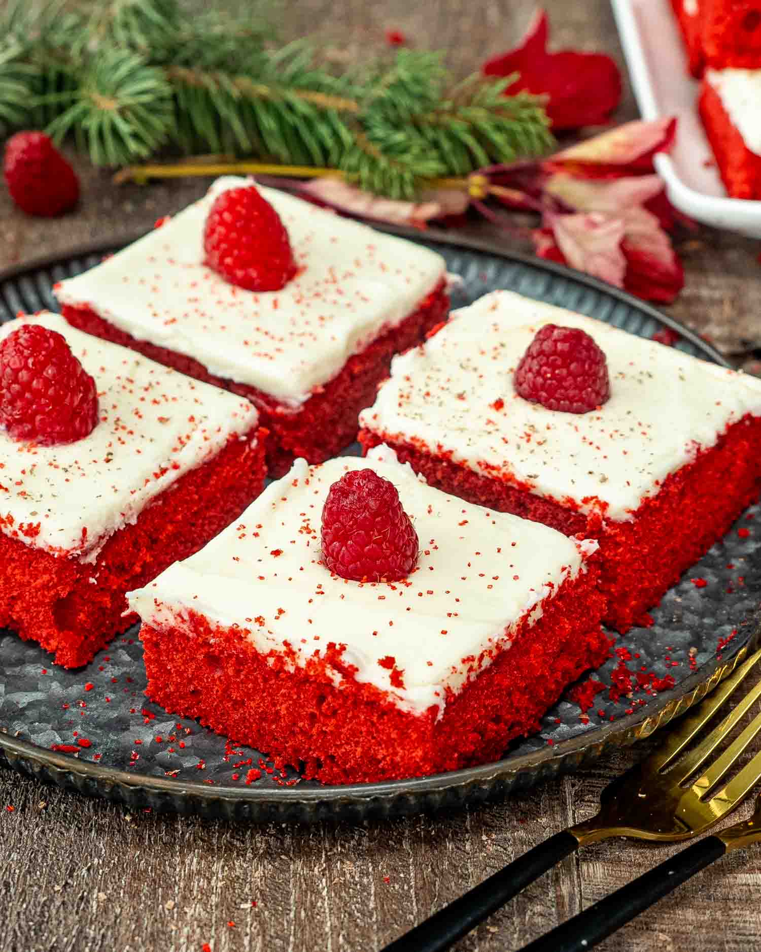 4 slice of red velvet sheet cake topped with cream cheese frosting and each topped with a raspberry on a metal plate.