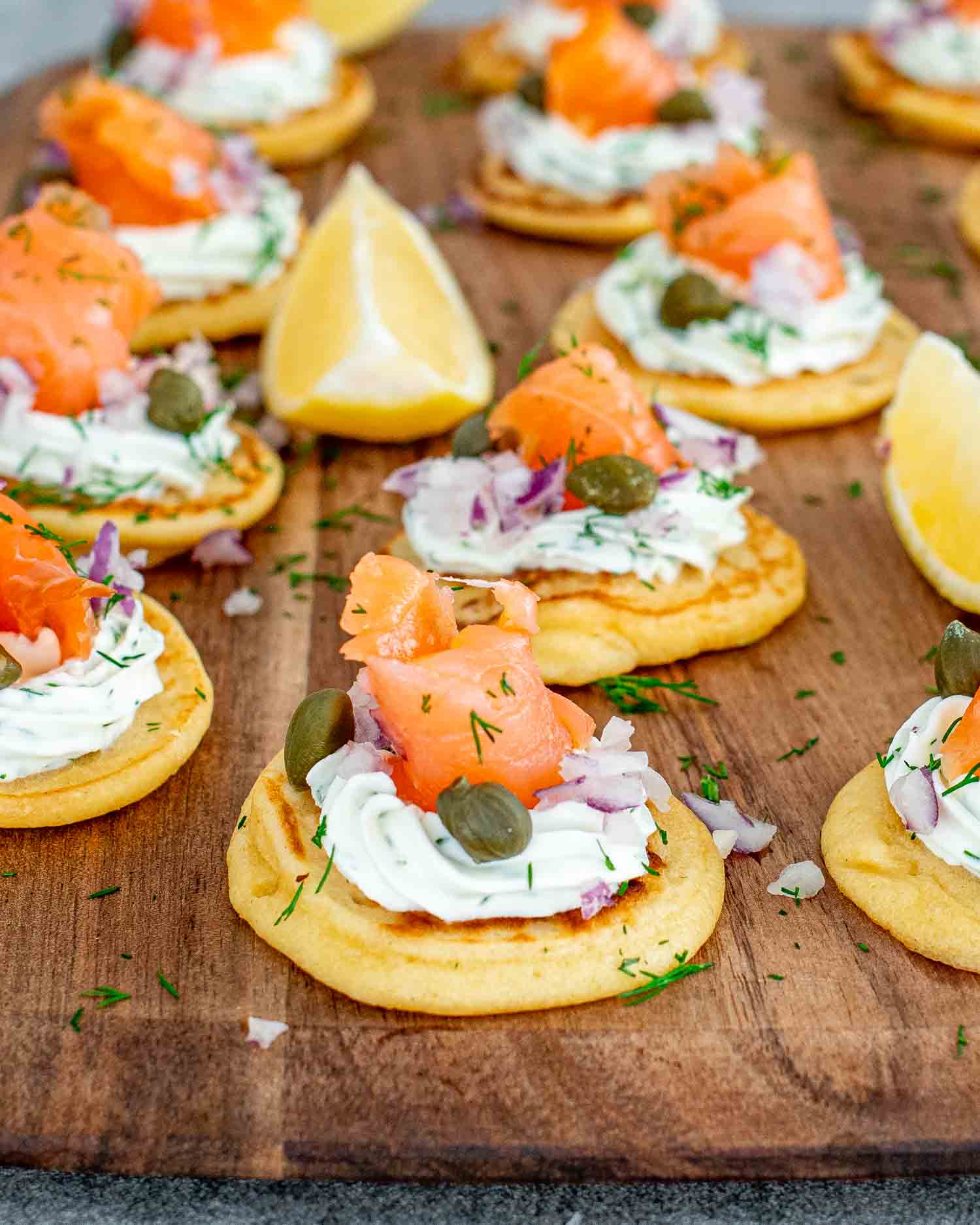 freshly made smoked salmon blinis on a cutting board garnished with lemon wedges.