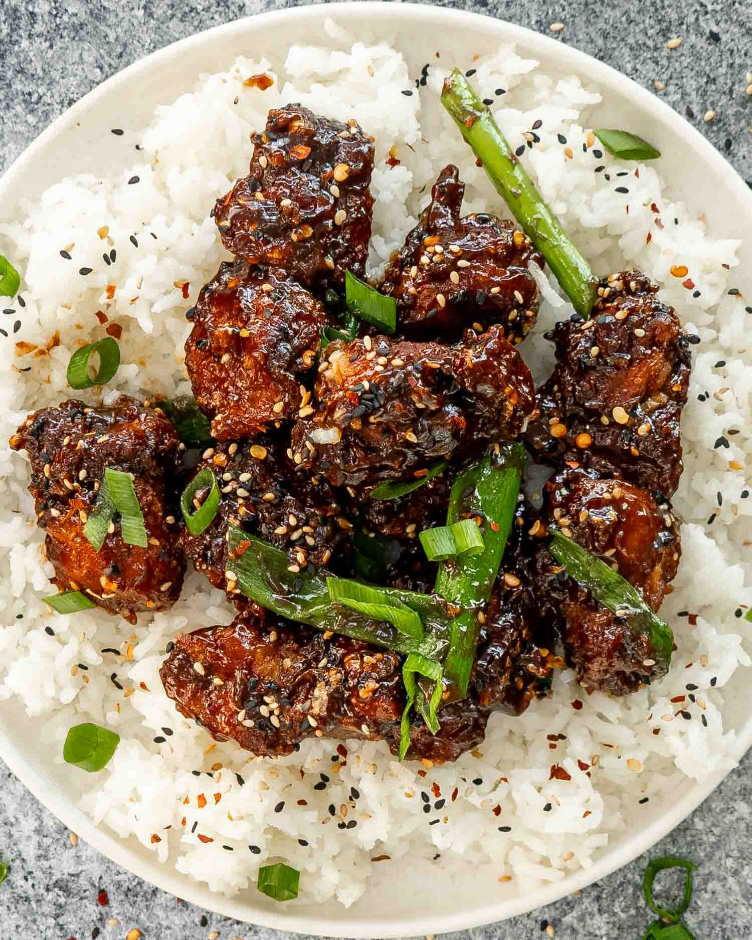 freshly made general tso's chicken garnished with green onions and sesame seeds over a rice in a white plate.