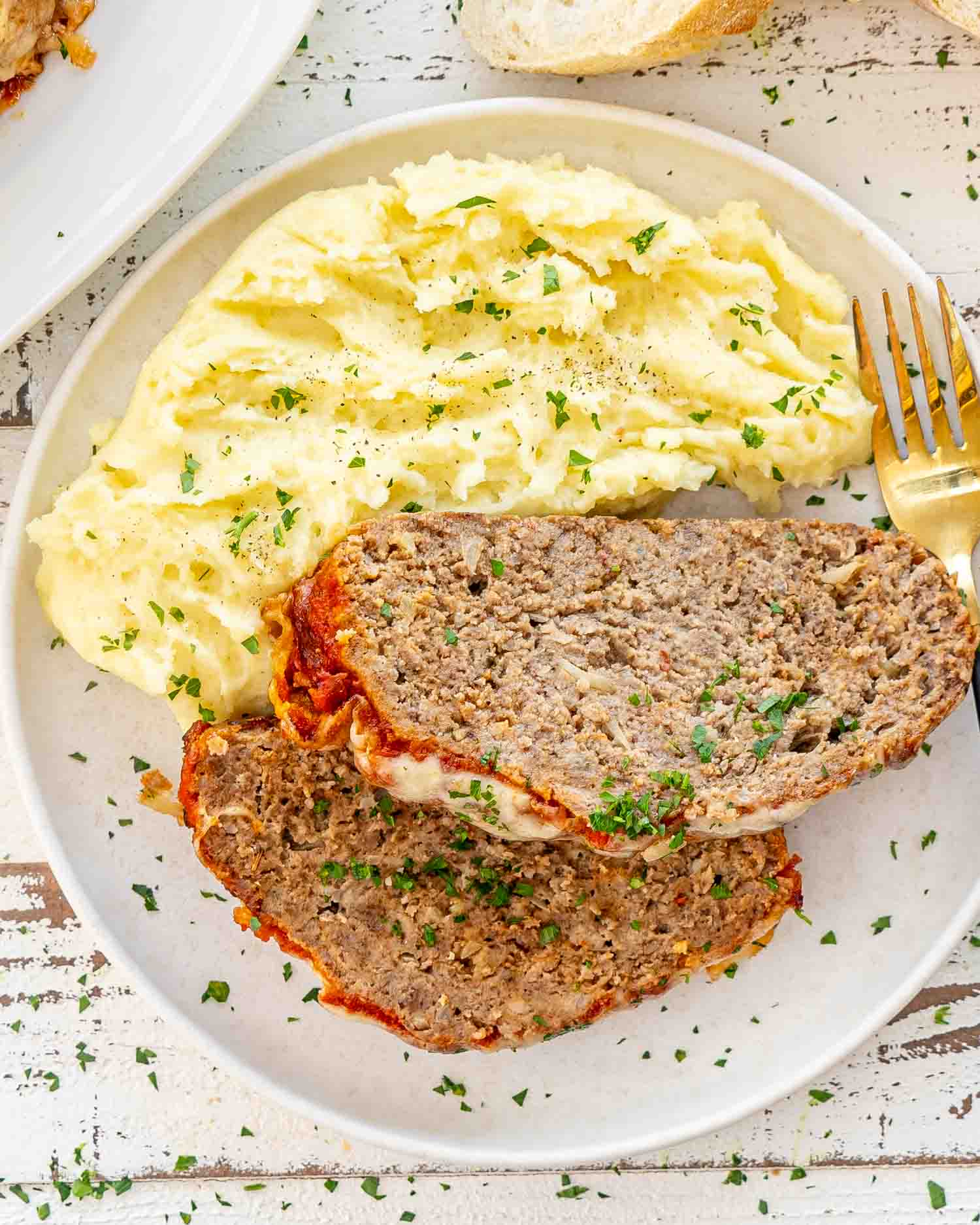 2 slices of italian meatloaf along some fluffy mashed potatoes garnished with fresh parsley on a white plate.