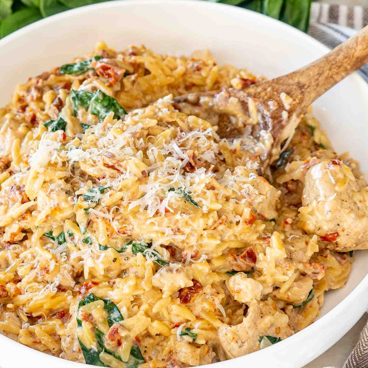 slow cooker creamy tuscan chicken orzo in a white serving bowl with a wooden spoon inside.