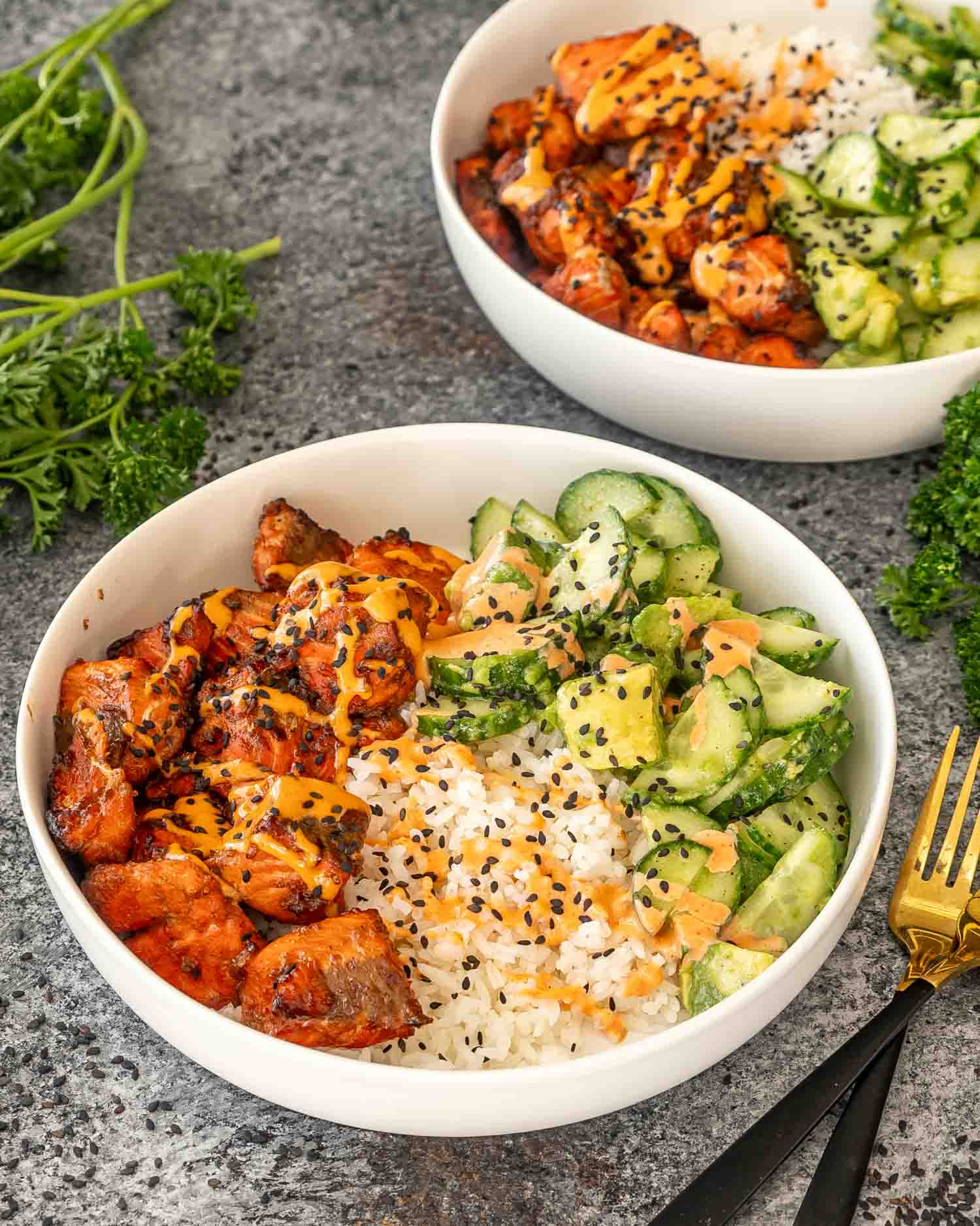 teriyaki salmon bites that were made in the air fryer, in a white bowl with steamed rice, cucumber avocado salad and a sriracha mayo.