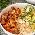 teriyaki salmon bites that were made in the air fryer, in a white bowl with steamed rice, cucumber avocado salad and a sriracha mayo.