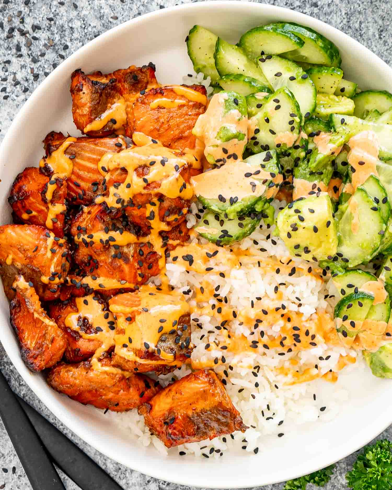 teriyaki salmon bites that were made in the air fryer, in a white bowl with steamed rice, cucumber avocado salad and a sriracha mayo.