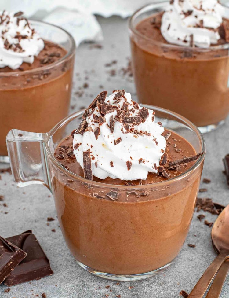 A glass of chocolate mousse topped with whipped cream and chocolate shavings on a gray surface.
