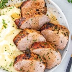 slices of herb crusted pork tenderloin along some mashed potatoes on a white plate.