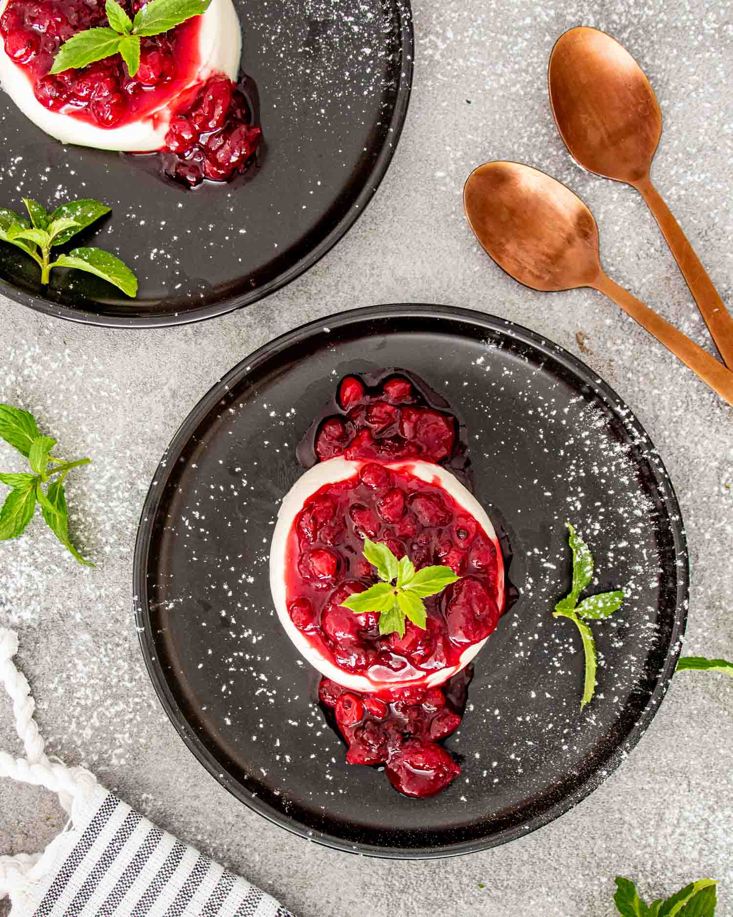 Panna cotta topped with vibrant red berry sauce and a sprig of mint on a dark plate.