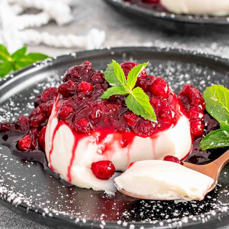Panna cotta topped with vibrant red berry sauce and a sprig of mint on a dark plate.
