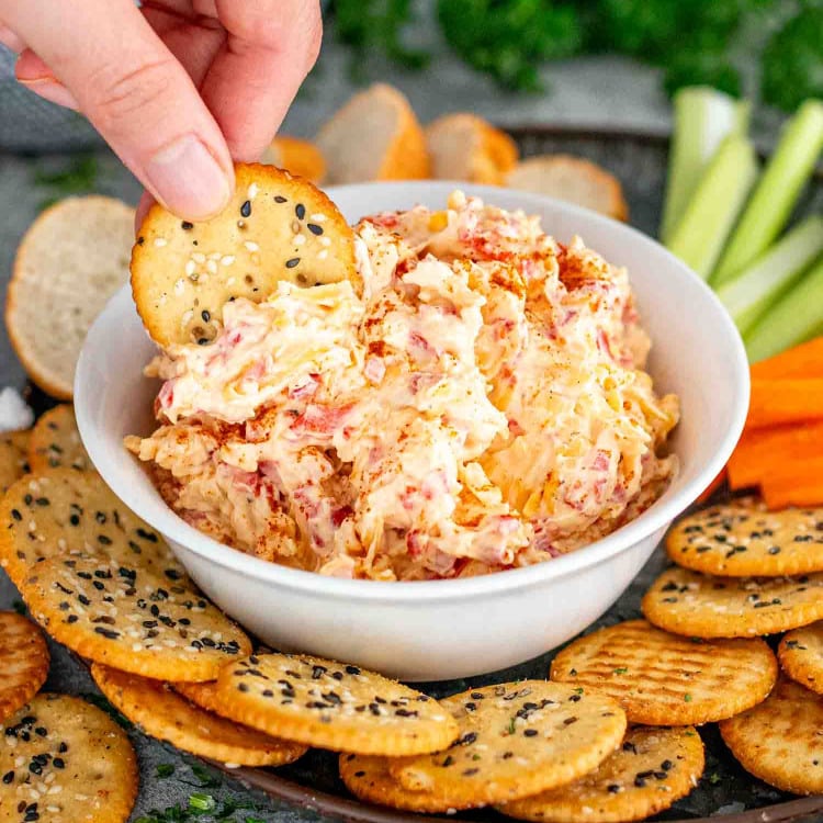 pimento cheese in a white bowl along some crackers, celery and carrot sticks.