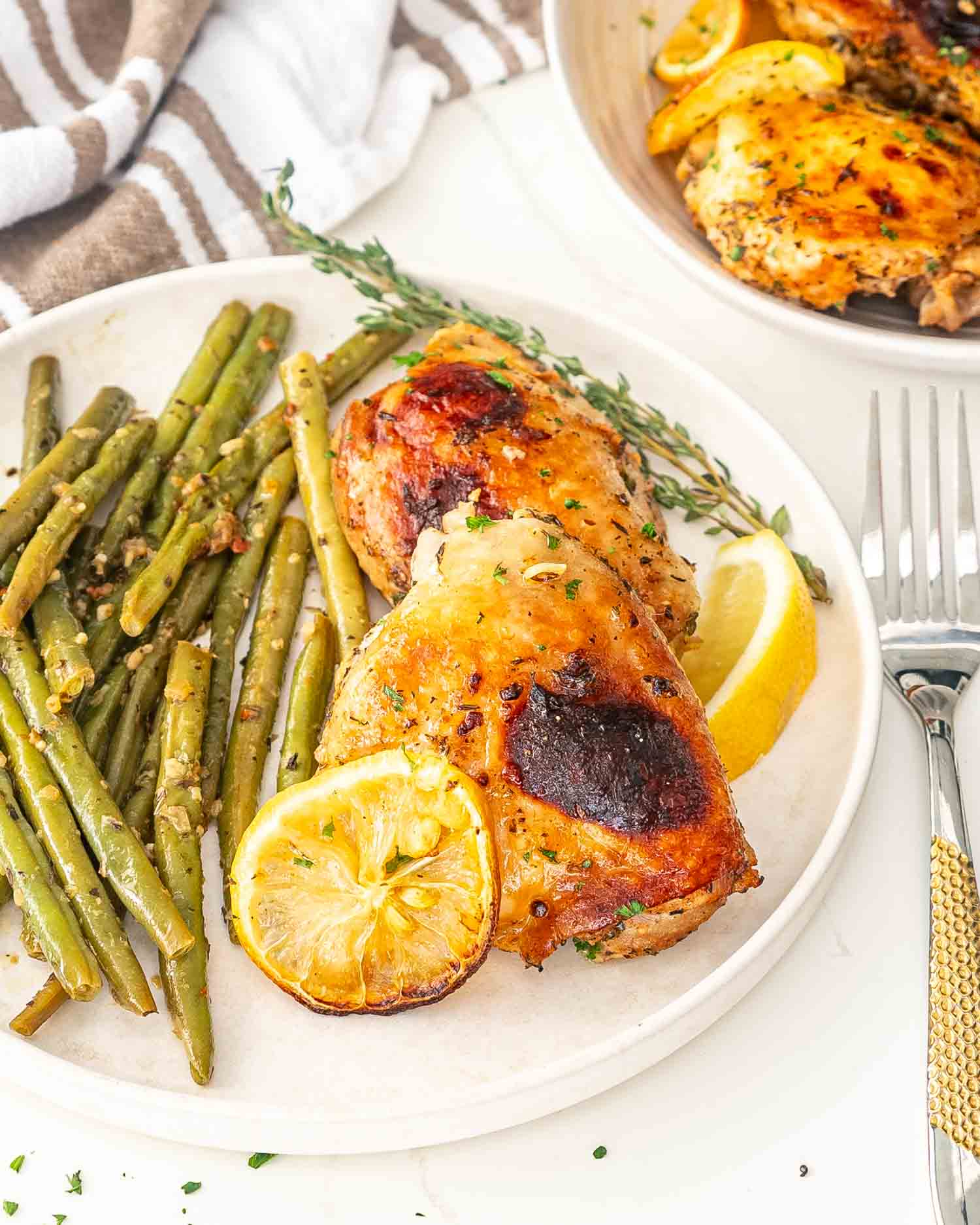 two baked lemon chicken thighs on a white plate along a side of green beans.