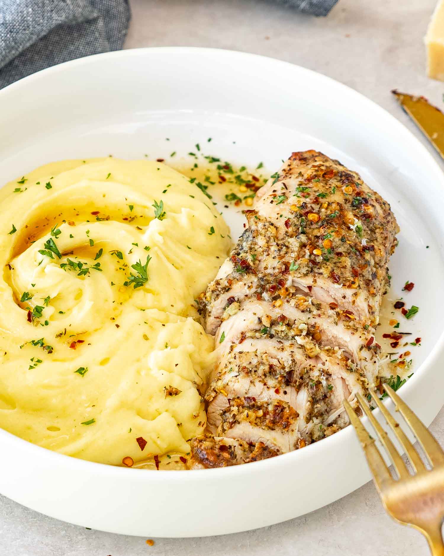 a parmesan herb chicken breast on a plate with mashed potatoes garnished with parsley.