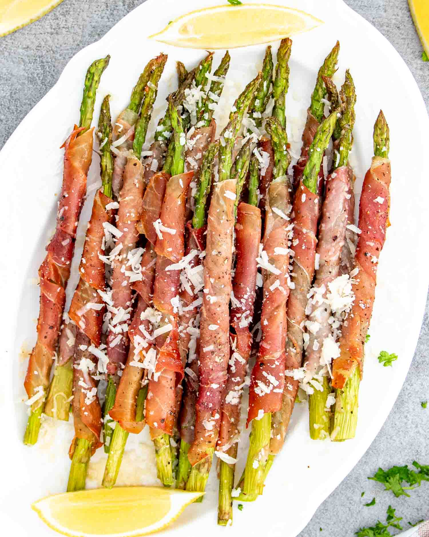 prosciutto wrapped asparagus on a serving platter garnished with parmesan cheese and lemon wedges.